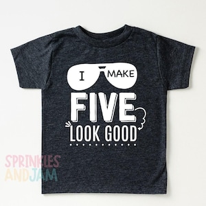 May include: A dark heather gray short-sleeve t-shirt featuring a white graphic of sunglasses and the text I MAKE FIVE LOOK GOOD. The design includes a small decorative swirl and a dotted line underneath the text, centered on the chest area.
