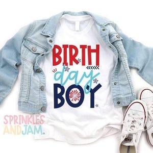 May include: A white t-shirt featuring the red, light blue, and navy blue text BIRTH day BOY with star and firework graphics, layered under a light blue denim jacket, accompanied by a pair of white canvas sneakers with white laces and rubber soles.