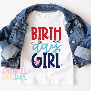 May include: A white t-shirt featuring the text BIRTH day GIRL in red, light blue, and dark blue, accented with small stars and a firework graphic, layered under a blue denim jacket with silver buttons and cuffed long sleeves.