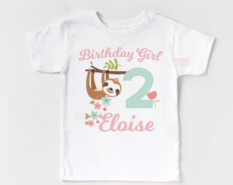 May include: A white short-sleeved t-shirt featuring a graphic of a brown sloth hanging from a branch, a large mint green number 2, and pink floral accents. The text reads Birthday Girl and Eloise in a pink script font with a small logo.