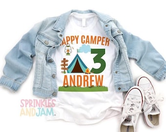 May include: A white t-shirt featuring the text HAPPY CAMPER and ANDREW in orange, a blue tent, a lantern, a campfire, and a large green number 3. The shirt is layered under a light blue denim jacket and paired with white canvas sneakers.