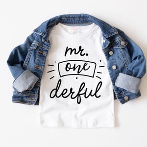 May include: A white short-sleeved t-shirt featuring the black text "mr. one derful" layered under a blue denim jacket with metal buttons. The shirt has a round neckline and the text is centered with decorative lines surrounding the word one.