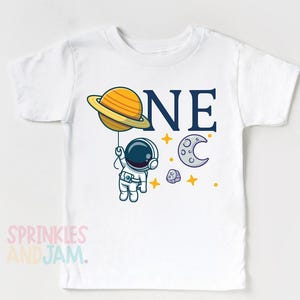 May include: A white short-sleeved t-shirt featuring a graphic of an astronaut holding a yellow planet like a balloon. The text "ONE" is printed in dark blue, with a crescent moon, small stars, and a rock graphic surrounding the astronaut.