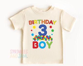 May include: A cream-colored short-sleeve t-shirt featuring a graphic print with the text BIRTHDAY in multi-colored letters, a large blue number 3, and the word BOY in red, blue, and green letters, accented by colorful polka dots and sprinkles.