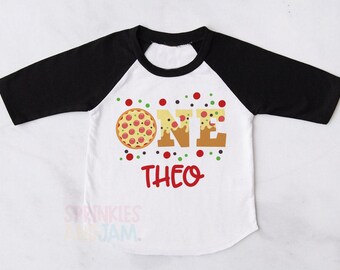 May include: A white raglan t-shirt with black three-quarter length sleeves. The front features a graphic of a pizza replacing the letter O in the word ONE, with the name THEO printed in red below. Small red and green dots surround the text.