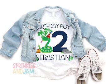 May include: A white t-shirt featuring a graphic of a green snake wearing a red party hat, a large navy blue number 2, and the text BIRTHDAY BOY SEBASTIAN. The shirt is layered under a light blue denim jacket, accompanied by white canvas sneakers.