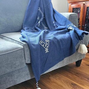 May include: A blue throw blanket with white embroidered designs and tassels. The blanket is draped over a gray couch.