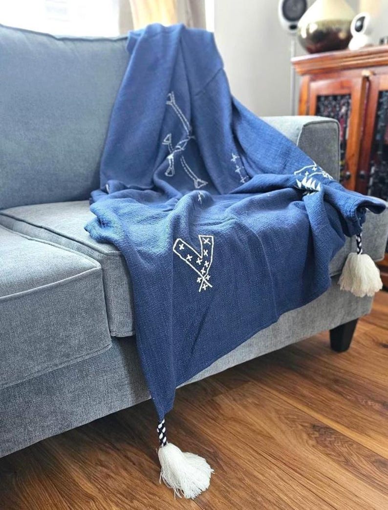 May include: A blue throw blanket with white embroidered designs and tassels. The blanket is draped over a gray couch.
