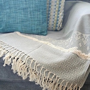 Puede incluir: Una manta gris con un patrón geométrico y flecos. La manta está colocada sobre un sofá gris con dos almohadas, una azul verdosa y otra con un patrón blanco y verde.