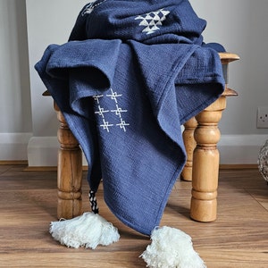 May include: A navy blue throw blanket with white geometric patterns and tassels. The blanket is draped over a wooden stool.