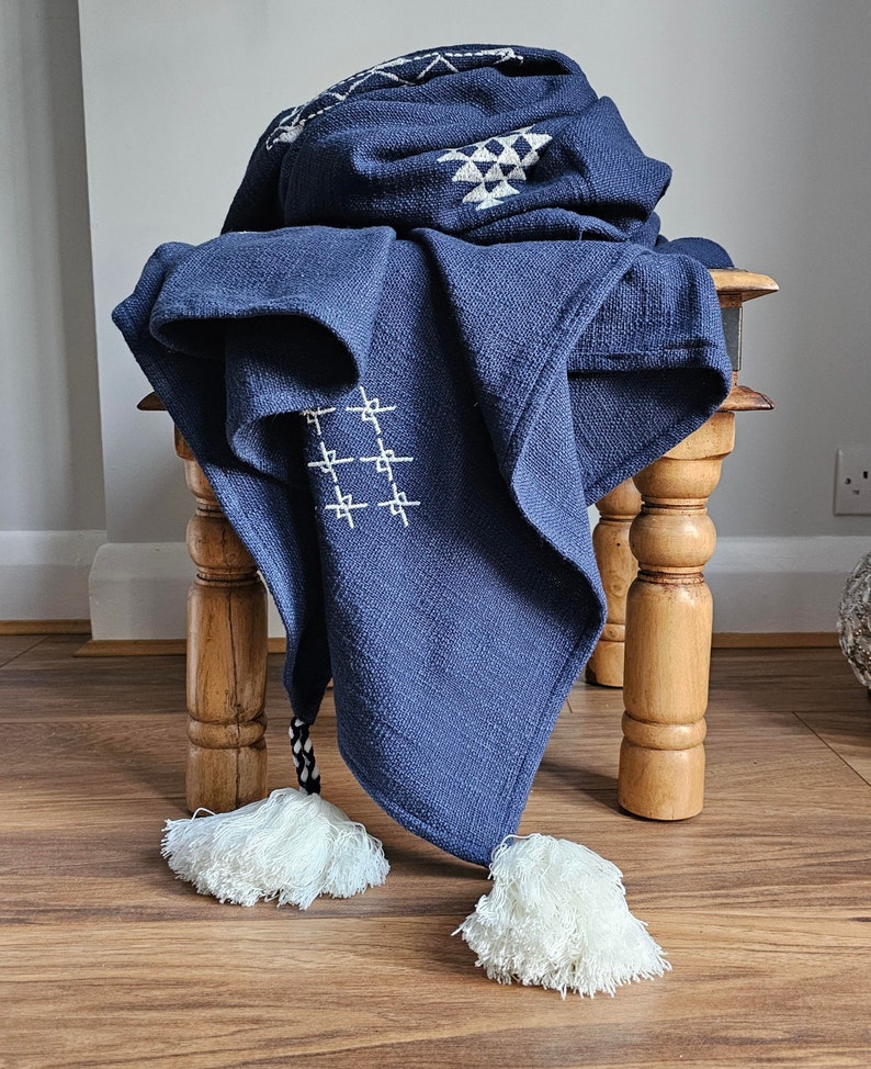 May include: A navy blue throw blanket with white geometric patterns and tassels. The blanket is draped over a wooden stool.