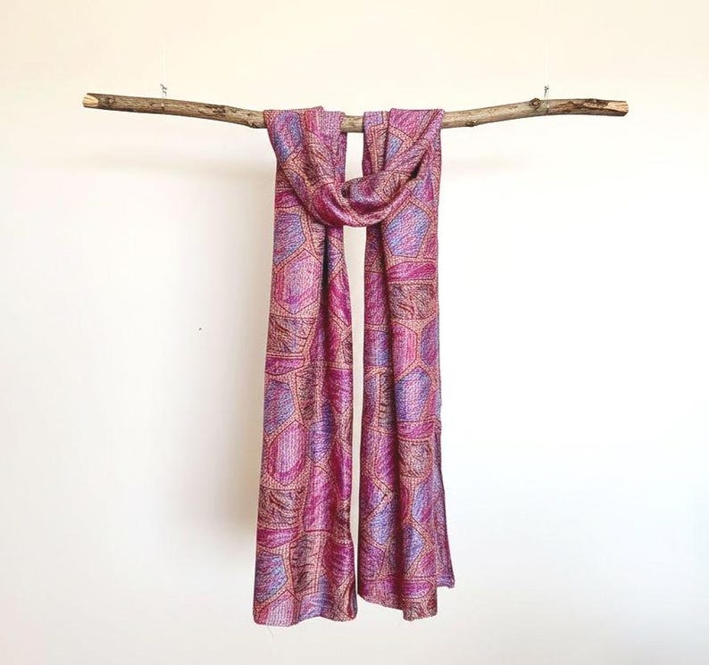 Colourful abstract print pink silk scarf draped over a rustic wooden branch suspended from the ceiling against a plain background