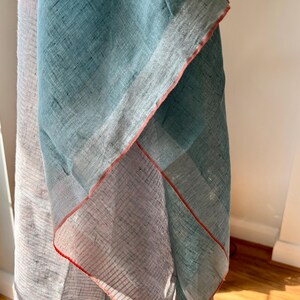 May include: A light blue and white linen scarf with a red trim. The scarf is folded in half and the edges are visible.