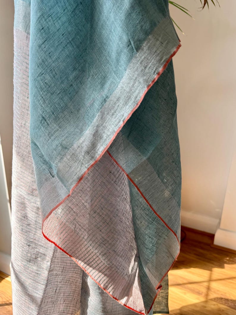 May include: A light blue and white linen scarf with a red trim. The scarf is folded in half and the edges are visible.