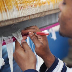 Indian artisan sat in front of handloom carefully separating threads whilst weaving linen kaftan