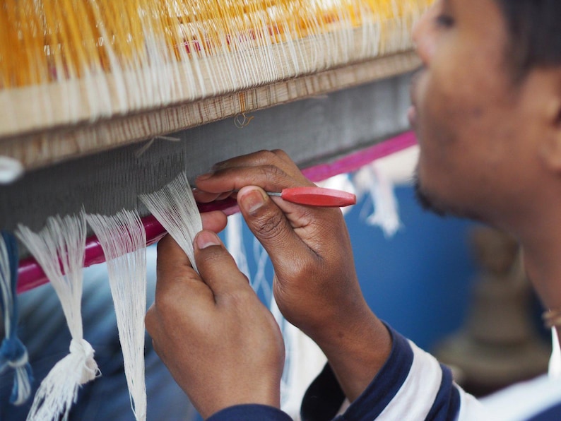 Indian artisan sat in front of handloom carefully separating threads whilst weaving linen kaftan
