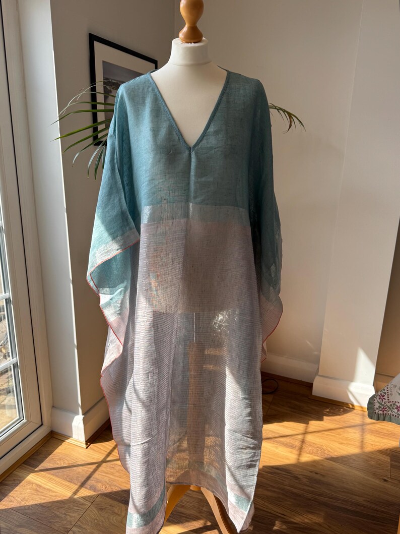 May include: A long, flowing, light blue and gray linen kaftan with a V-neckline. The kaftan has a loose, comfortable fit and is perfect for warm weather.