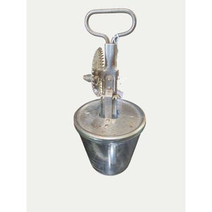 May include: Vintage metal butter churn with a handle and gear mechanism. The churn has a cylindrical container with a lid and a handle on top. The gears and handle are made of metal. The churn is likely used for making butter.