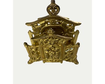 May include: A gold-colored metal napkin holder with an ornate design. The holder has a decorative top and intricate details throughout.
