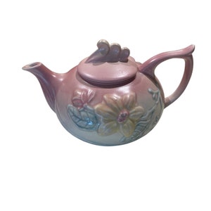 May include: A pink glazed ceramic teapot with a floral design. The teapot has a lid with a decorative knob and a spout. The flowers are in shades of yellow, pink, and green.