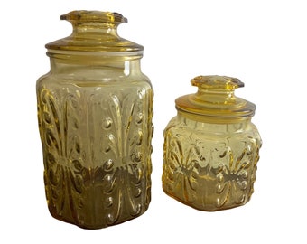 Pode incluir: Dois frascos de vidro âmbar com desenhos florais em relevo. O frasco maior é mais alto e tem uma base mais larga do que o frasco menor.