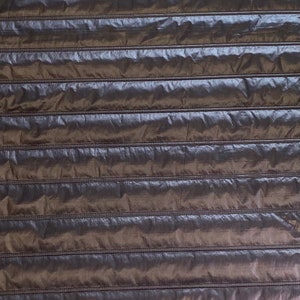 Op de afbeelding: Een close-up van een donkerbruine, gestikte stof met horizontale naden. De stof heeft een licht glanzende, bijna iriserende uitstraling.