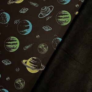May include: Dark brown fabric featuring a repeating pattern of planets, comets, and Saturn-like rings in blue, green, and yellow. A solid black fabric is folded over the patterned fabric.