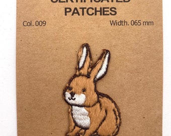 Könnte beinhalten: Ein braunes gesticktes Hasen-Patch mit weißen Akzenten. Das Patch befindet sich auf einer braunen Karte mit dem Text "Art. No. XQS22-009-065 CERTIFICATED PATCHES Col. 009 Width. 0,65 cm 50% Polyester - 50% Rayon - by Quality Trimmings".