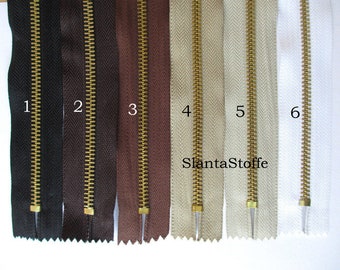 Könnte beinhalten: Sechs verschiedene Reißverschlussbänder mit goldenen Metallreißverschlüssen. Die Bänder sind in folgenden Farben: Schwarz, Braun, Braun, Beige, Beige und Weiß.