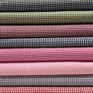 Könnte beinhalten: Ein Stapel aus zehn gefalteten Stoffballen mit einem klassischen Vichy-Karo-Muster in verschiedenen Farben, darunter Limettengrün, Schwarz, Oliv, Dunkelrot, Marineblau, Magenta, Hellrosa, Grau, Hellrot und Hellblau, die alle horizontal angeordnet sind.