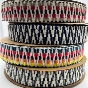 Puede incluir: Una pila de cuatro carretes de cinta decorativa. Las cintas presentan patrones tejidos en varios colores, incluyendo rojo, negro, blanco, azul marino y amarillo. Los carretes son de cartón.