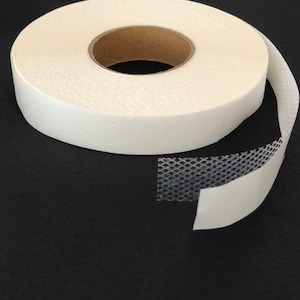 Puede incluir: Un rollo de cinta adhesiva de doble cara blanca con un respaldo de malla negra. La cinta está sobre una superficie negra.