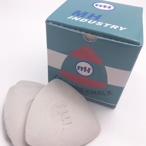 May include: A box of white tailor's chalk in the shape of a triangle. The box is blue and white with the text "MH INDUSTRY" and a logo of a rocket ship with the letters "MH" inside. The box is sitting on a white surface.