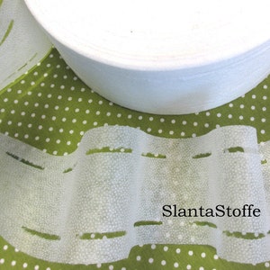 Könnte beinhalten: Weißer Stoffbesatz mit gepunkteter Textur und einer Reihe gleichmäßig verteilter Schlitze. Der Besatz ist auf einer weißen Spule aufgerollt. Der Text "SlantaStoffe" ist auf dem Besatz sichtbar.