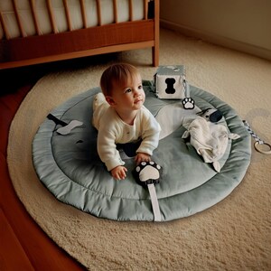 Peut inclure: Un tapis d'éveil rond vert sauge au motif de chien, doté de jouets en peluche attachés, dont un cube souple avec un imprimé d'os, un hochet en forme de patte et un jouet en tissu texturé. Le tapis présente une bordure matelassée et des tons neutres.