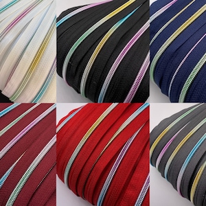Könnte beinhalten: Sortiment von Reißverschlussbändern in verschiedenen Farben, darunter Weiß, Schwarz, Marineblau, Burgunderrot, Rot und Grau. Jedes Band ist mit einem goldenen, silbernen und türkisfarbenen Besatz versehen.