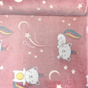 Könnte beinhalten: Eine weiche, rosafarbene Plüschdecke mit einem wiederkehrenden Muster aus weißen Cartoon-Katzen mit regenbogenfarben gestreiften Einhorn-Hörnern. Das Design enthält weiße Mondsicheln und kleine fünfzackige Sterne auf der Stoffoberfläche.