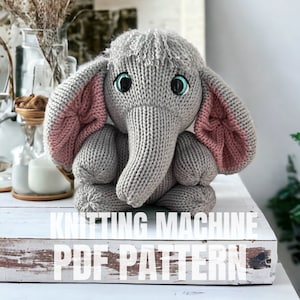 May include: Knitted gray elephant toy with pink ears. The toy has blue eyes and is sitting on a white wooden surface. The text "Knitting Machine PDF Pattern" is written below the toy.