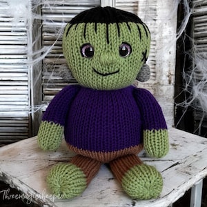 May include: A green knitted Frankenstein monster doll with a purple sweater, brown trousers, and sparkly pink eyes. The doll is sitting on a white wooden surface.