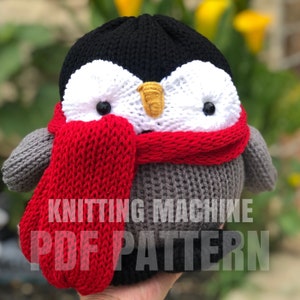 Op de afbeelding: Breimachine PDF-patroon voor een grijs-zwarte pinguïn met een rode sjaal. De pinguïn heeft witte ogen en een gele snavel.