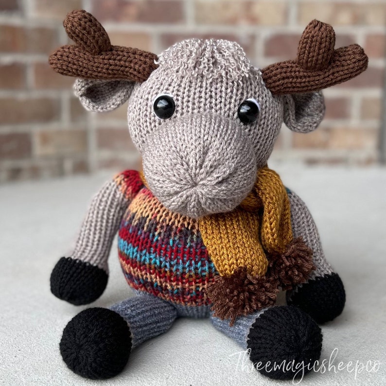 May include: A gray knitted moose wearing a colorful patterned sweater and a yellow scarf with brown pom-poms. The moose has large black eyes and brown antlers.