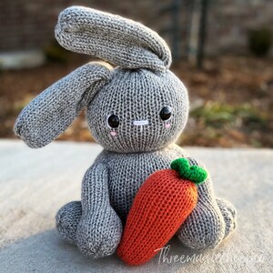 May include: A gray knitted bunny rabbit toy holding a knitted orange carrot with green leaves. The bunny has embroidered eyes and a white nose.