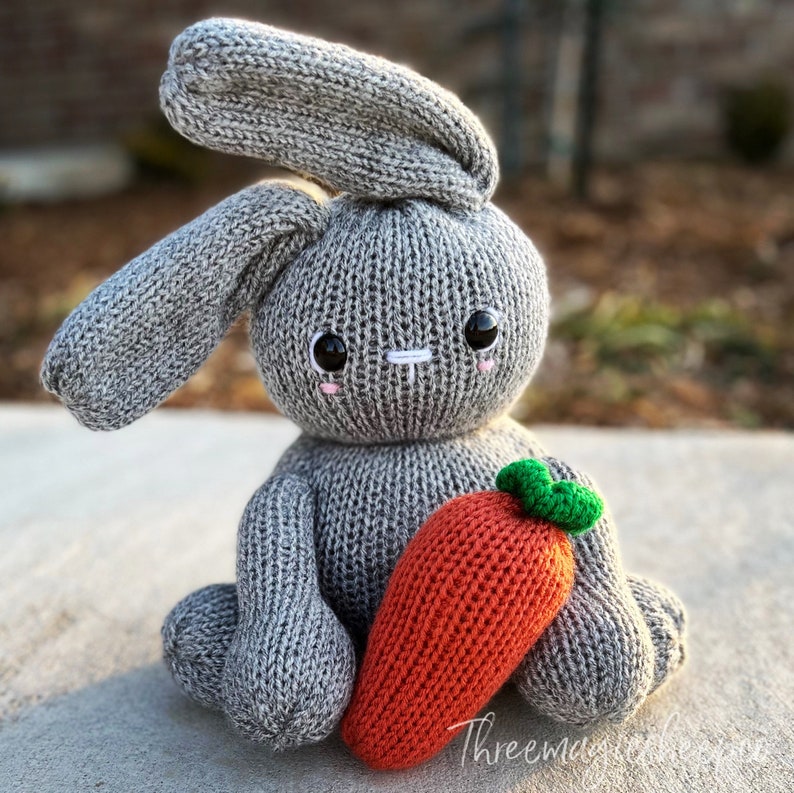 May include: A gray knitted bunny rabbit toy holding a knitted orange carrot with green leaves. The bunny has embroidered eyes and a white nose.