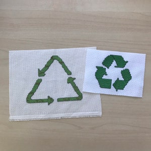 May include: Two white pieces of fabric with green embroidered recycling symbols. One symbol is a triangle with arrows pointing in a circle, the other is a traditional recycling symbol.