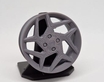 Op de afbeelding: Een grijs, 3D-geprint autowiel met een stervormig spaakontwerp. Het wiel is gemonteerd op een zwarte standaard, die de ingewikkelde details en textuur laat zien. De diameter van het wiel is ongeveer 15 cm.