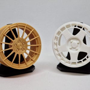 May include: Two model car wheels, one gold and one white, displayed on black stands. The gold wheel has a multi-spoke design, while the white wheel features a unique, open-faced pattern. Both are detailed and appear to be 3D-printed.