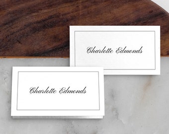 May include: Two white place cards with black borders and the text "Charlotte Edmonds" in cursive.