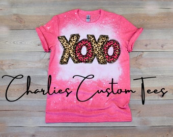 May include: A pink t-shirt with a bleached white area and the text "XOXO" in a leopard print design. The shirt also features the text "Charlies Custom Tees" in a cursive font.