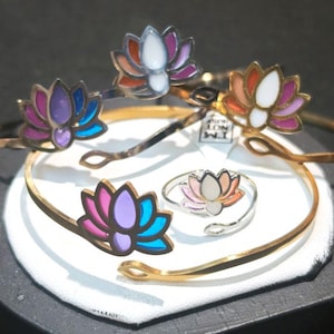 May include: A collection of gold-toned and silver-toned bracelets and a ring, each adorned with a colorful lotus flower design. The bracelets feature a hinged design, and the ring has an adjustable band. The lotus flowers are painted with shades of pink, purple, blue, and white.