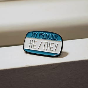 Puede incluir: Un pin de esmalte rectangular con forma de etiqueta de nombre con un borde azul y un centro blanco. El texto MY PRONOUNS est&aacute; escrito en azul en la parte superior, y el texto HE / THEY est&aacute; escrito en negro en el centro.